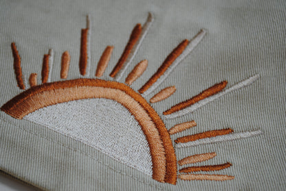 Close-up of an embroidered sun motif on a light sage green fabric, featuring warm brown and tan stitching with white rays.